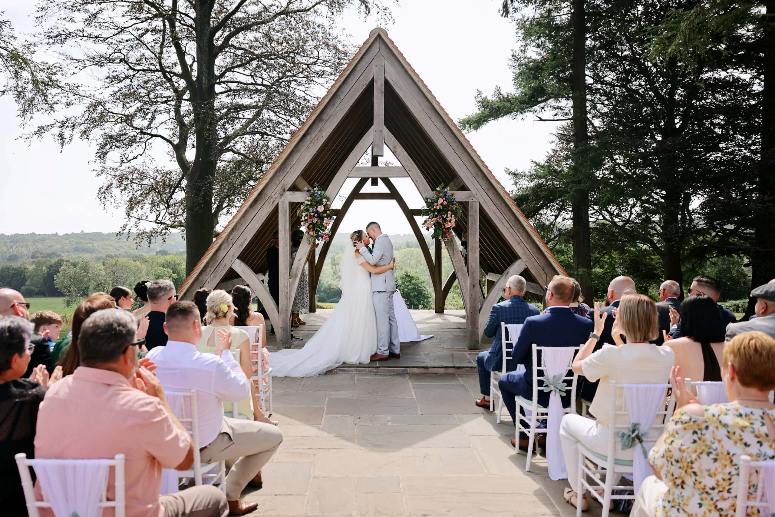 Elegant outdoor wedding ceremony at a modern wooden chapel surrounded by nature, featuring a bride and groom exchanging vows.